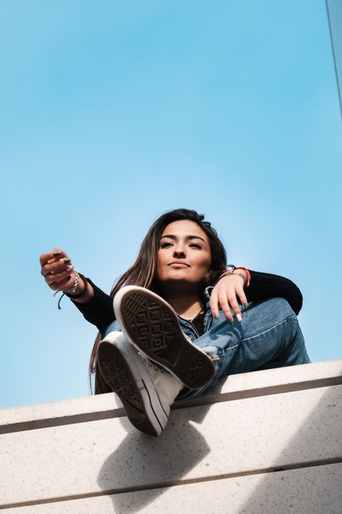 Free A fashionable woman in casual attire sitting on a ledge against a clear blue sky. Stock Photo