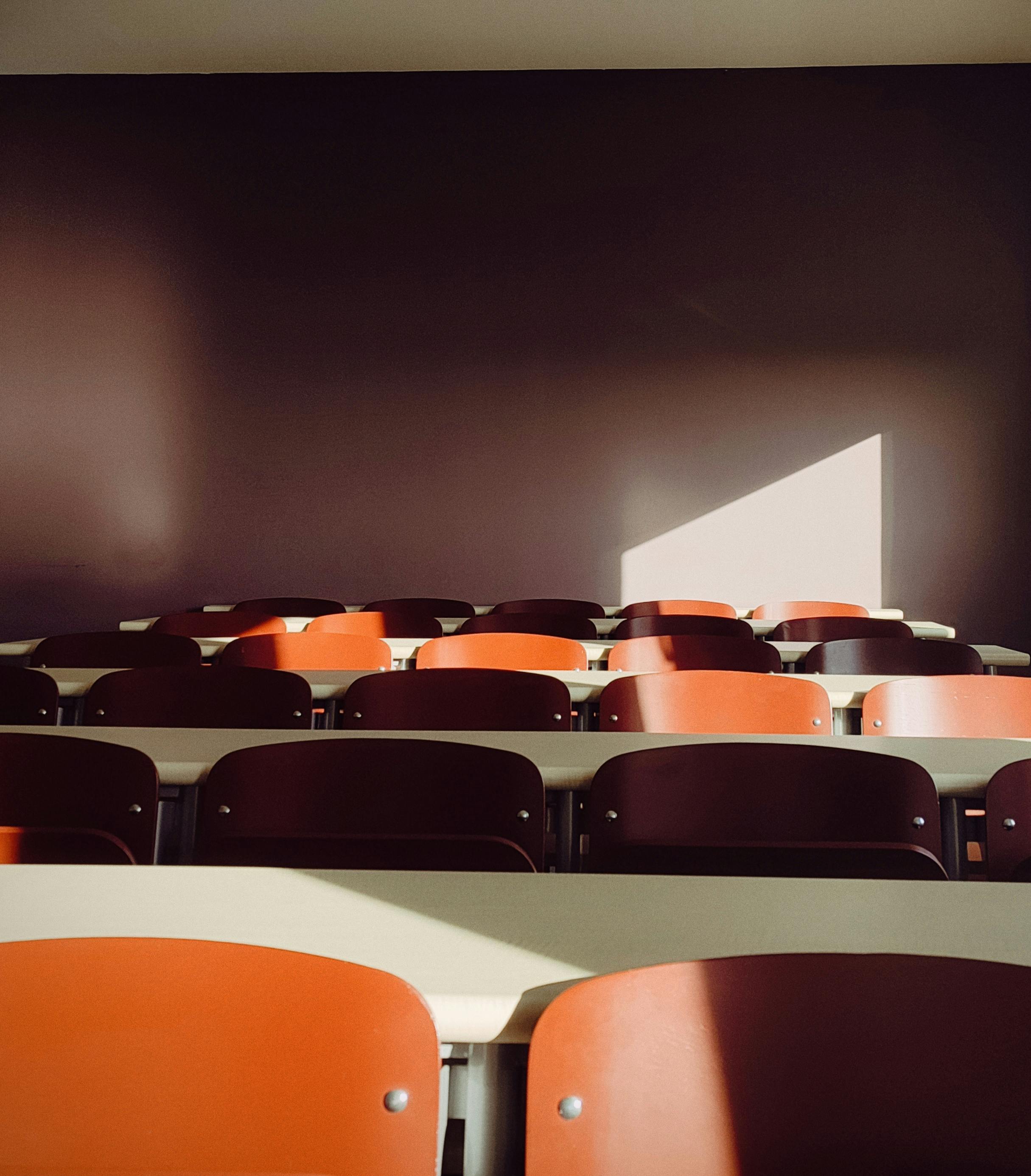 Free An empty classroom with sunlight casting shadows on red chairs, creating a dramatic effect. Stock Photo