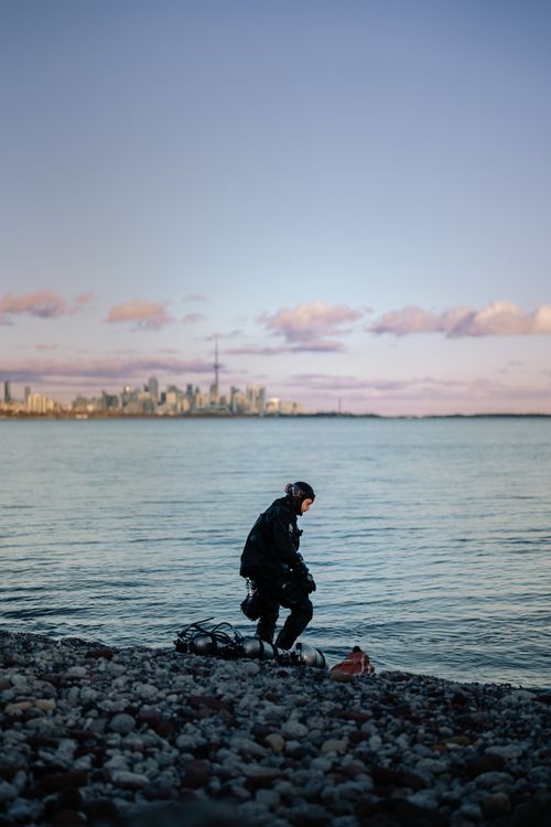 Free Scuba diver in gear on rocky shoreline with city skyline in the background during daylight. Stock Photo