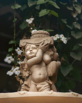 A charming cherub stone statue amidst lush green leaves and white flowers outdoors.
