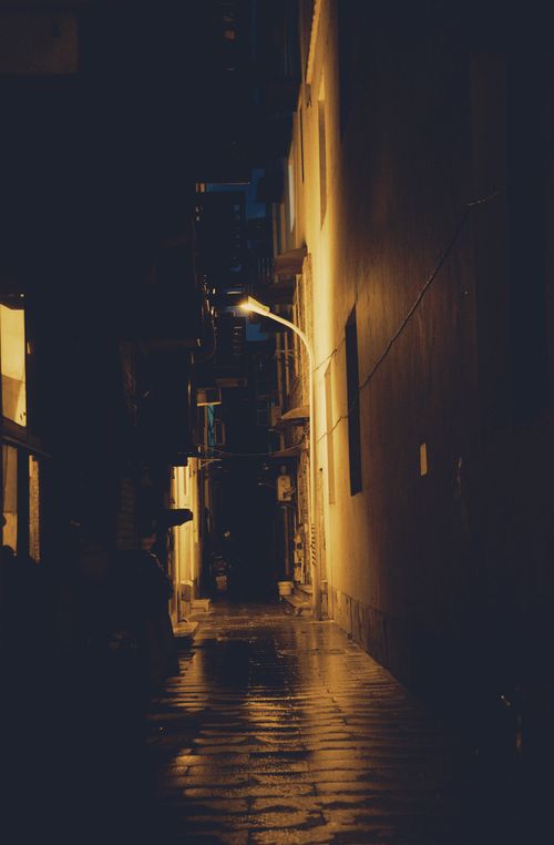 Free A dimly lit back alley at night features a wet pavement reflecting streetlamp light, evoking a moody urban setting. Stock Photo