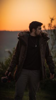 A man in a jacket holds a camera as the sun sets, capturing nature's serene moment.