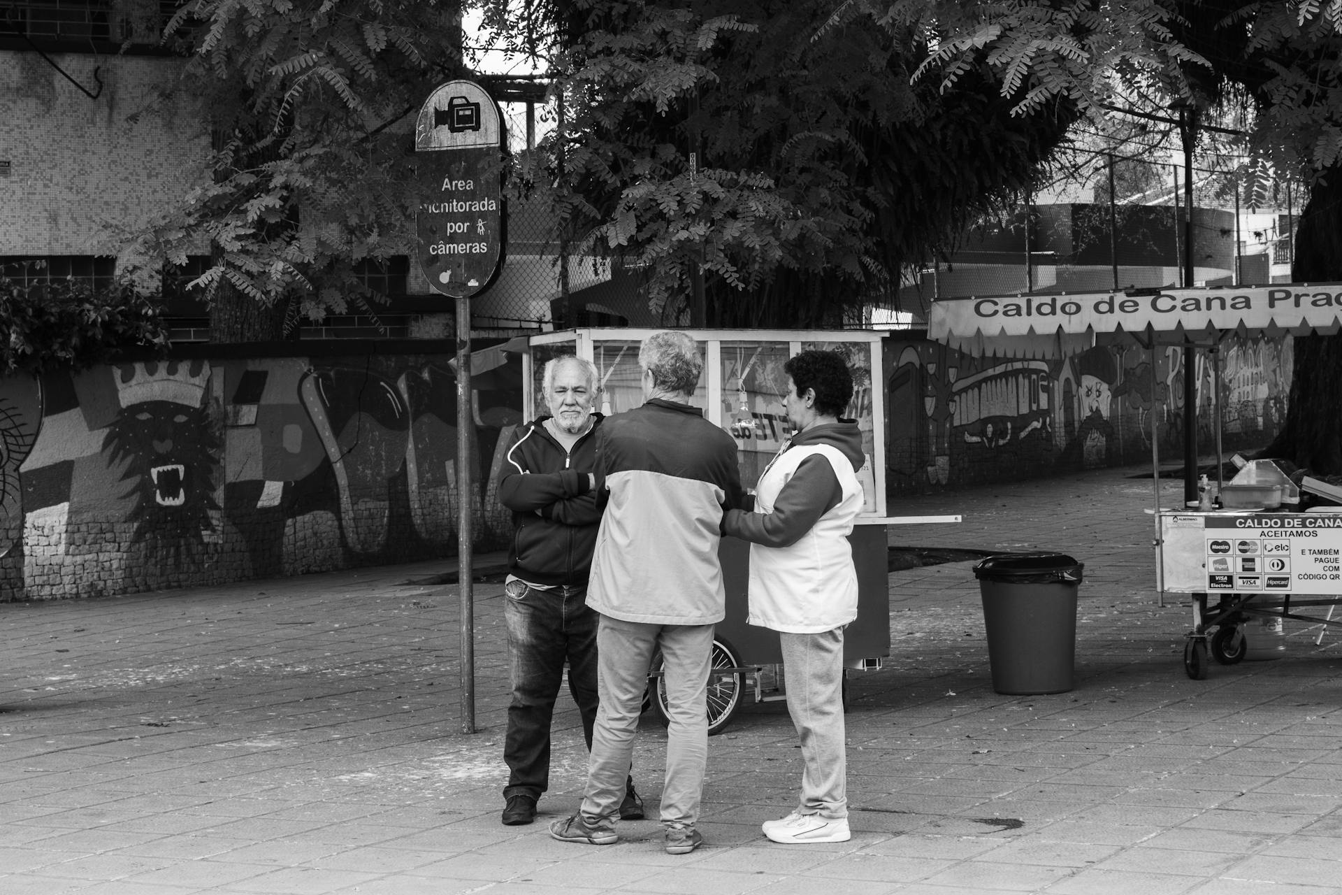 A black and white photo captures vendors and customers socializing at an urban street stall.