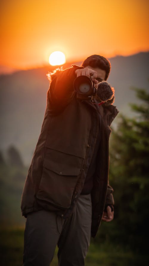 Man Holding a Camera in a Coniferous Forest During Sunset 