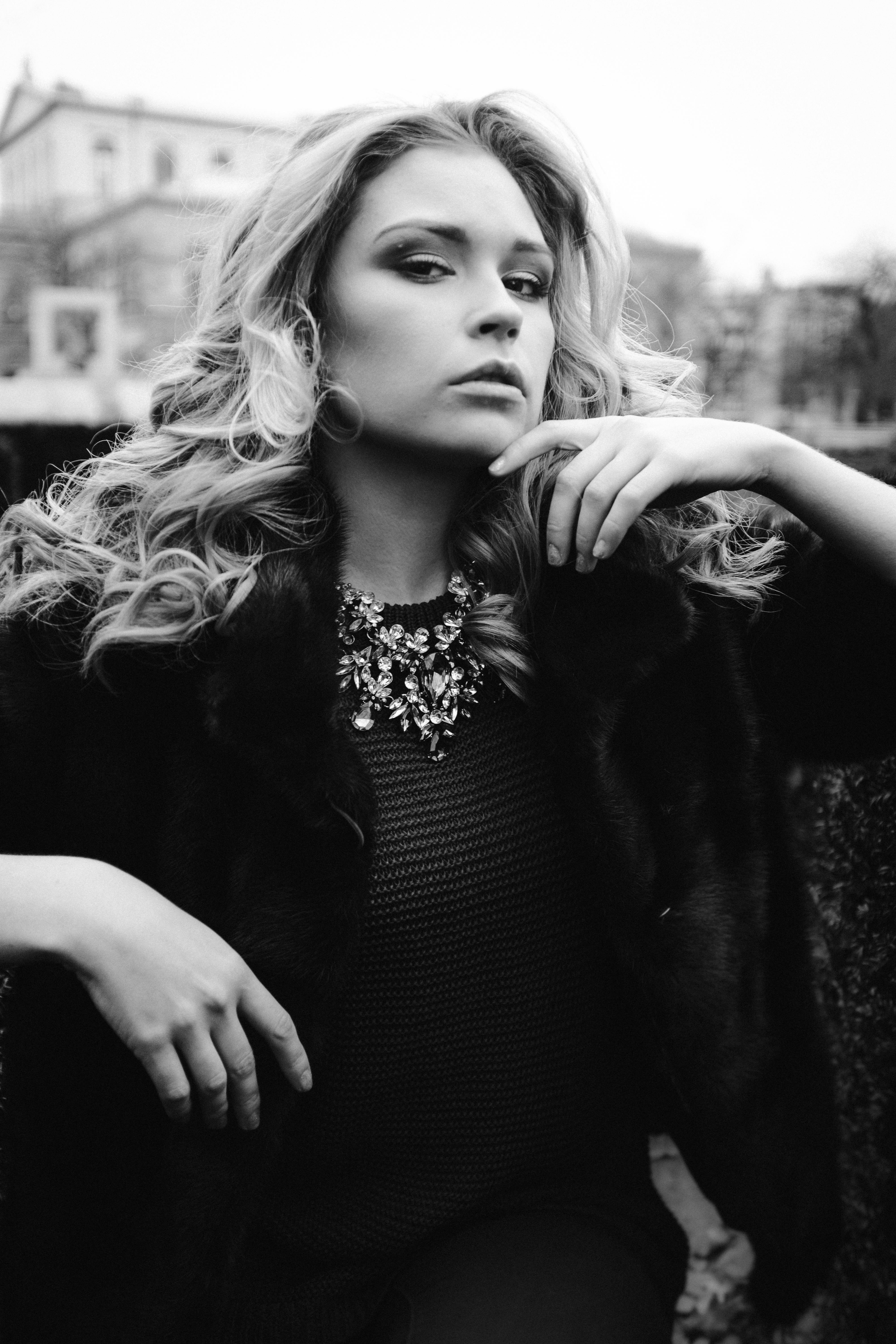 Black and white portrait of a woman in fur jacket and necklace, captured in urban environment.