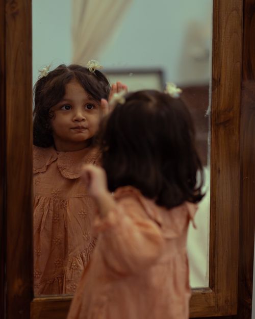 Free A young girl in a floral dress gazing at her reflection in a wooden-framed mirror. Stock Photo