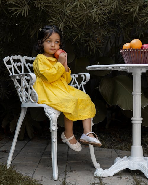 무료 사과를 들고 노란 드레스를 입고 의자에 앉아 있는 귀여운 소녀. 스톡 사진