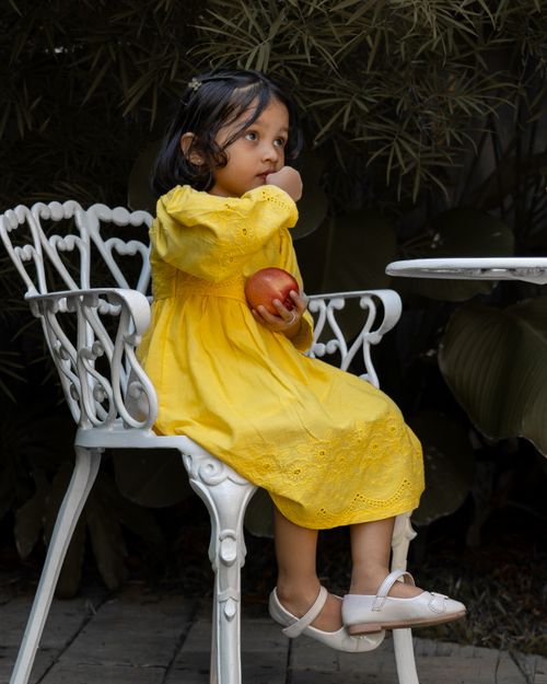 Free Adorable young girl in a yellow dress sitting on a white chair holding an apple. Stock Photo