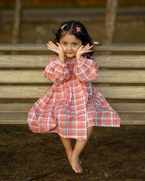 Free Adorable little girl in a plaid dress smiling on a bench outdoors. Stock Photo