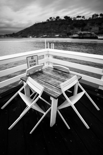 Black and white photo of a peaceful pier with empty chairs and a 'No Fishing' sign.