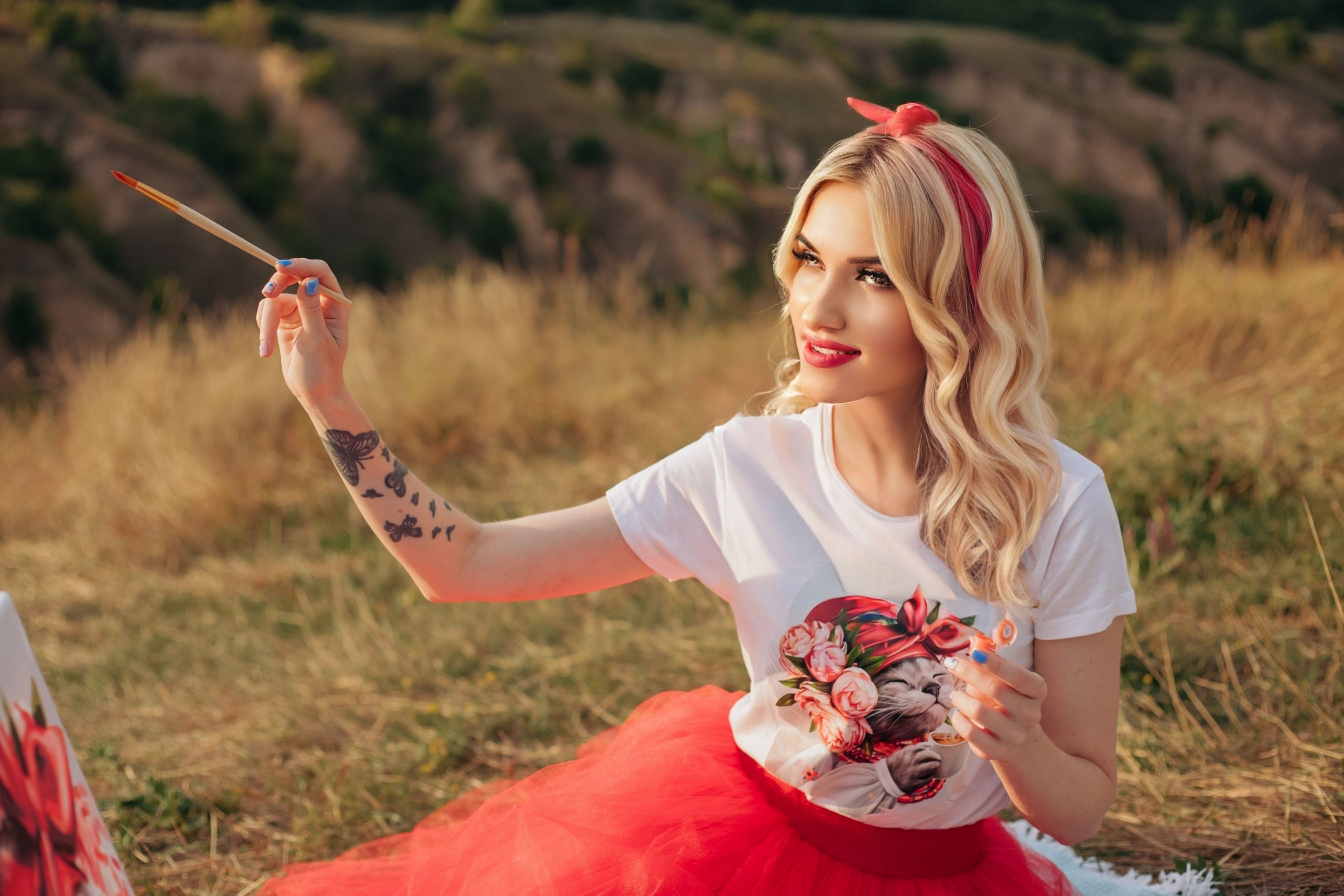 A fashionable young woman painting in a meadow, showcasing her artistic style with tattoos and vibrant clothing.