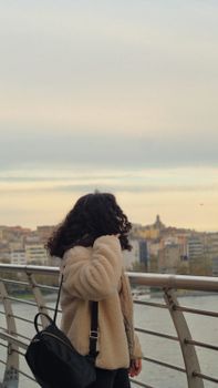 A woman with curly hair stands on a bridge, viewing the city at sunset.