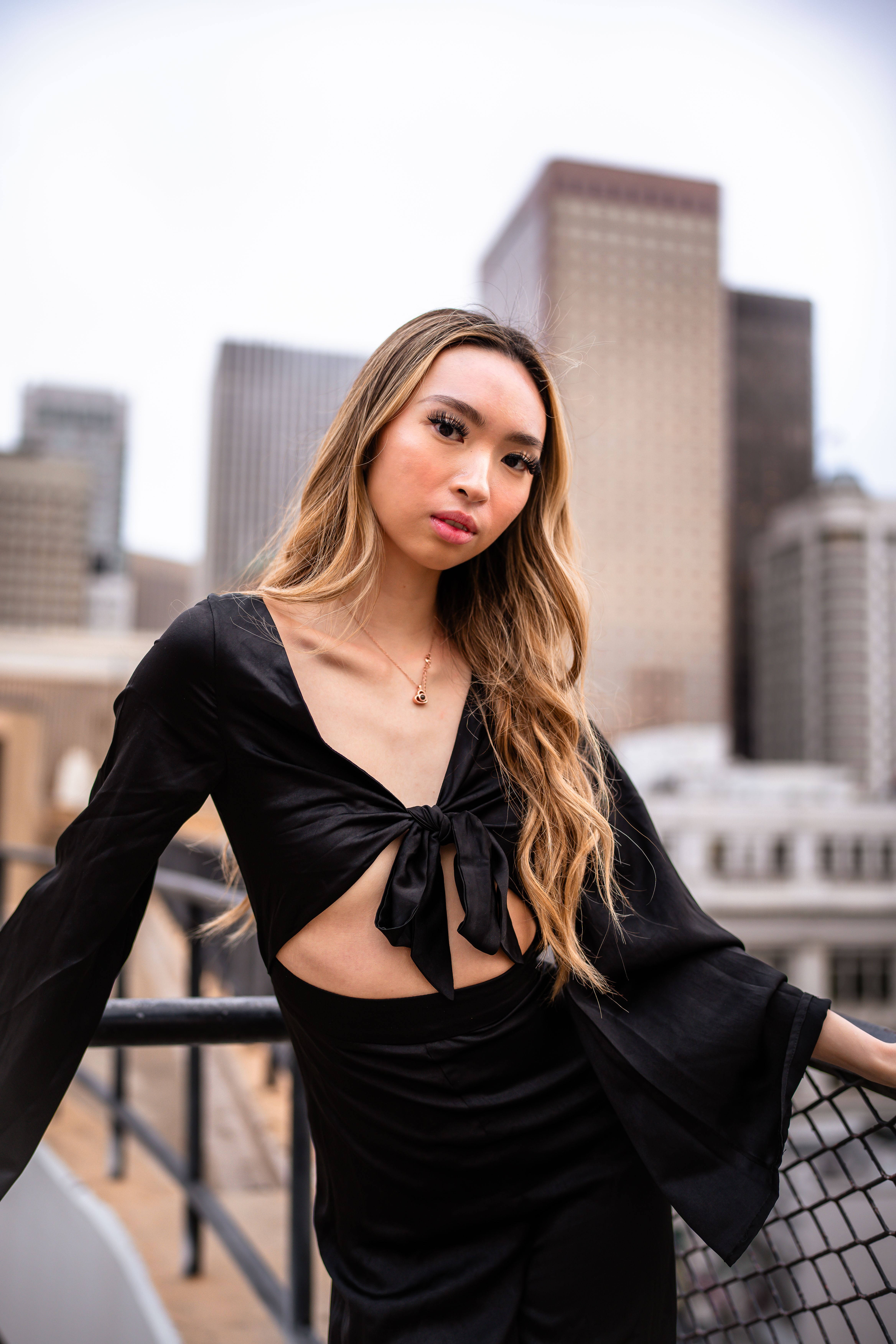 An elegant Asian woman poses in a trendy black dress on an urban rooftop, exuding fashion and glamour.