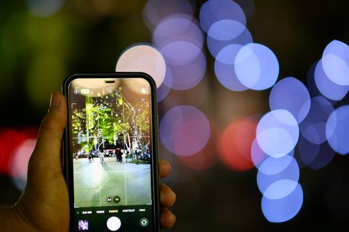 Free A close-up of a hand holding a smartphone capturing a night scene with bokeh lights. Stock Photo