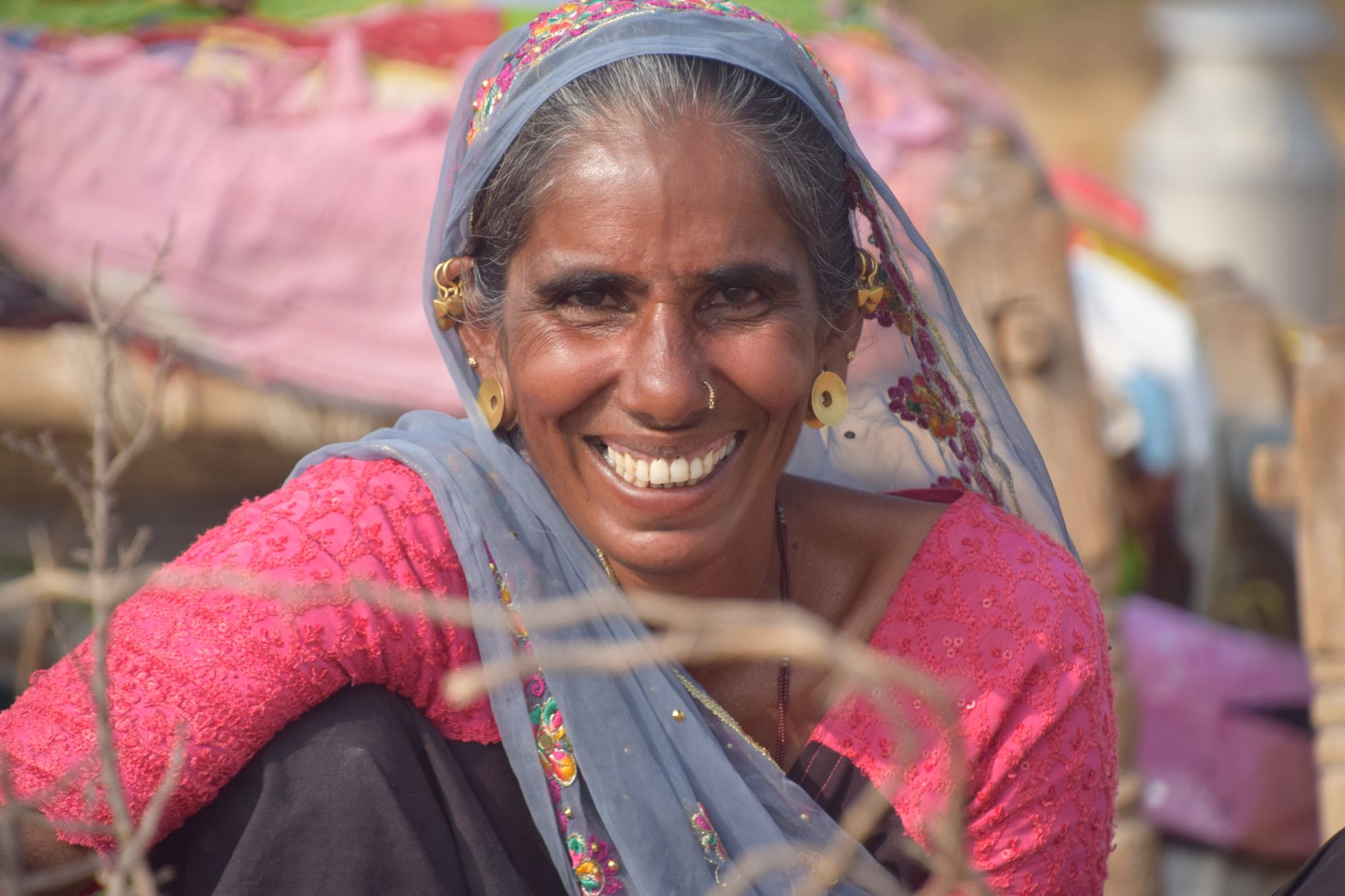 A South Asian woman smiles warmly, sitting outdoors in traditional clothing.