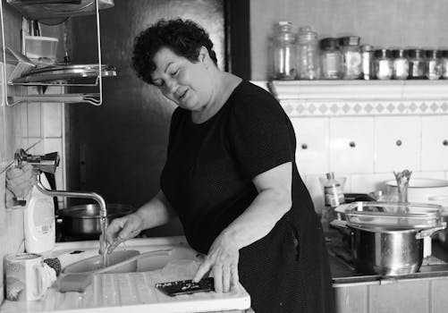 A candid black and white photo of a woman washing dishes in a cozy kitchen.