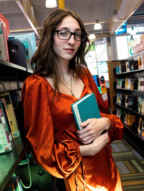 Young Woman in a Dress and Eyeglasses Standing in a Library and Holding a Book 