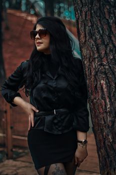Elegant woman in a black outfit standing by a tree, showcasing fashion and style.
