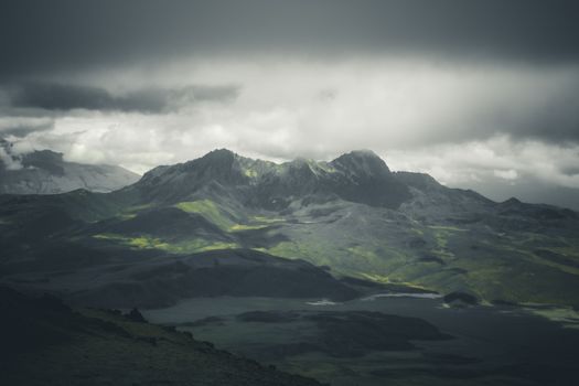 A moody mountain landscape with storm clouds casting shadows over green hills.