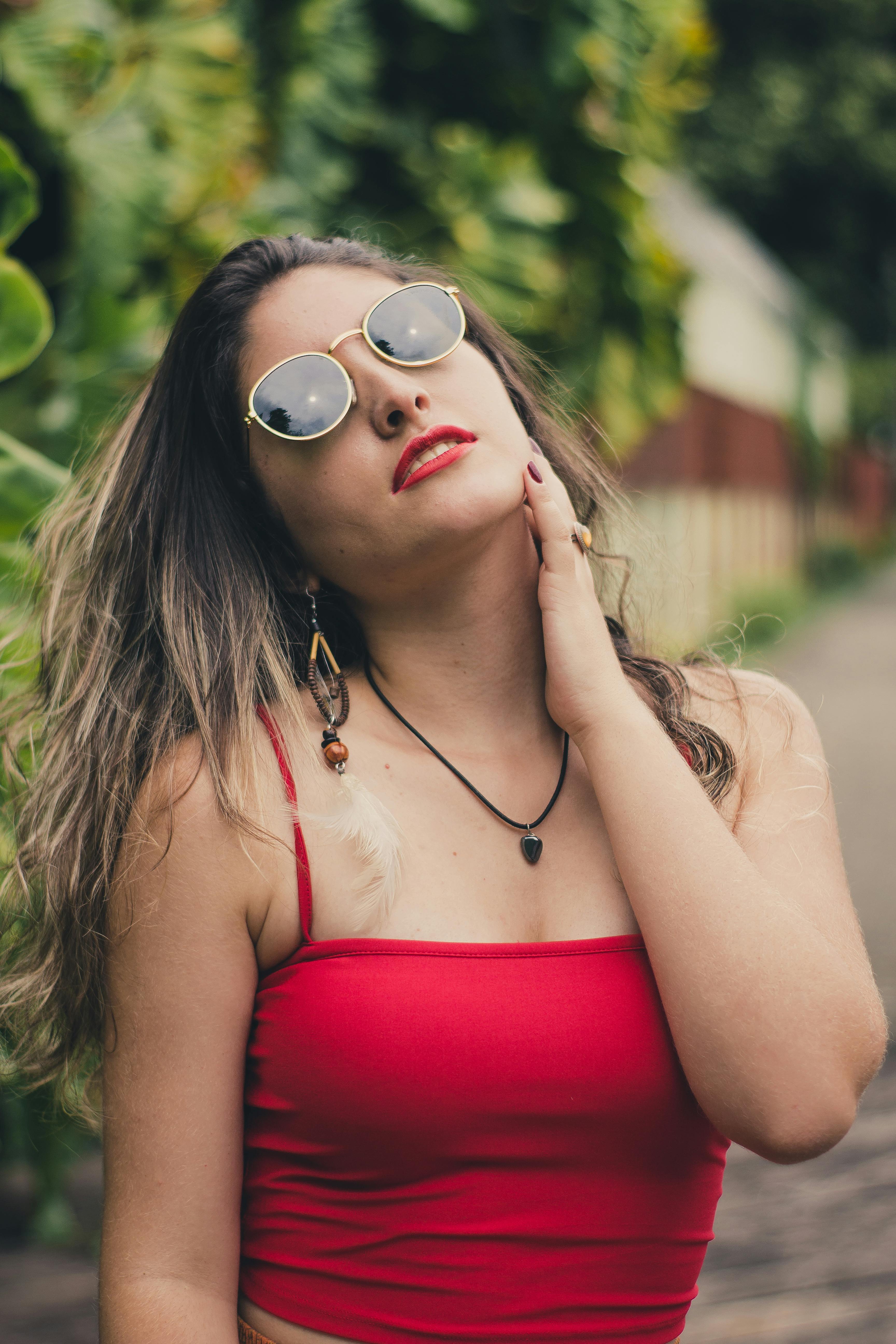 一位身穿红色上衣、戴着太阳镜的年轻女子在户外自信地摆着姿势，展现出独特的风格