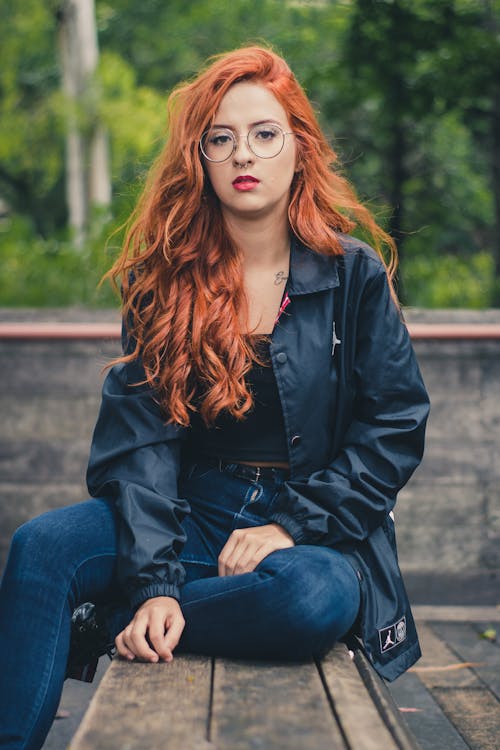 Mulher Com Jaqueta De Couro Preto Com Botões, Jeans Azul E óculos, Sentada Na Mesa De Madeira Marrom