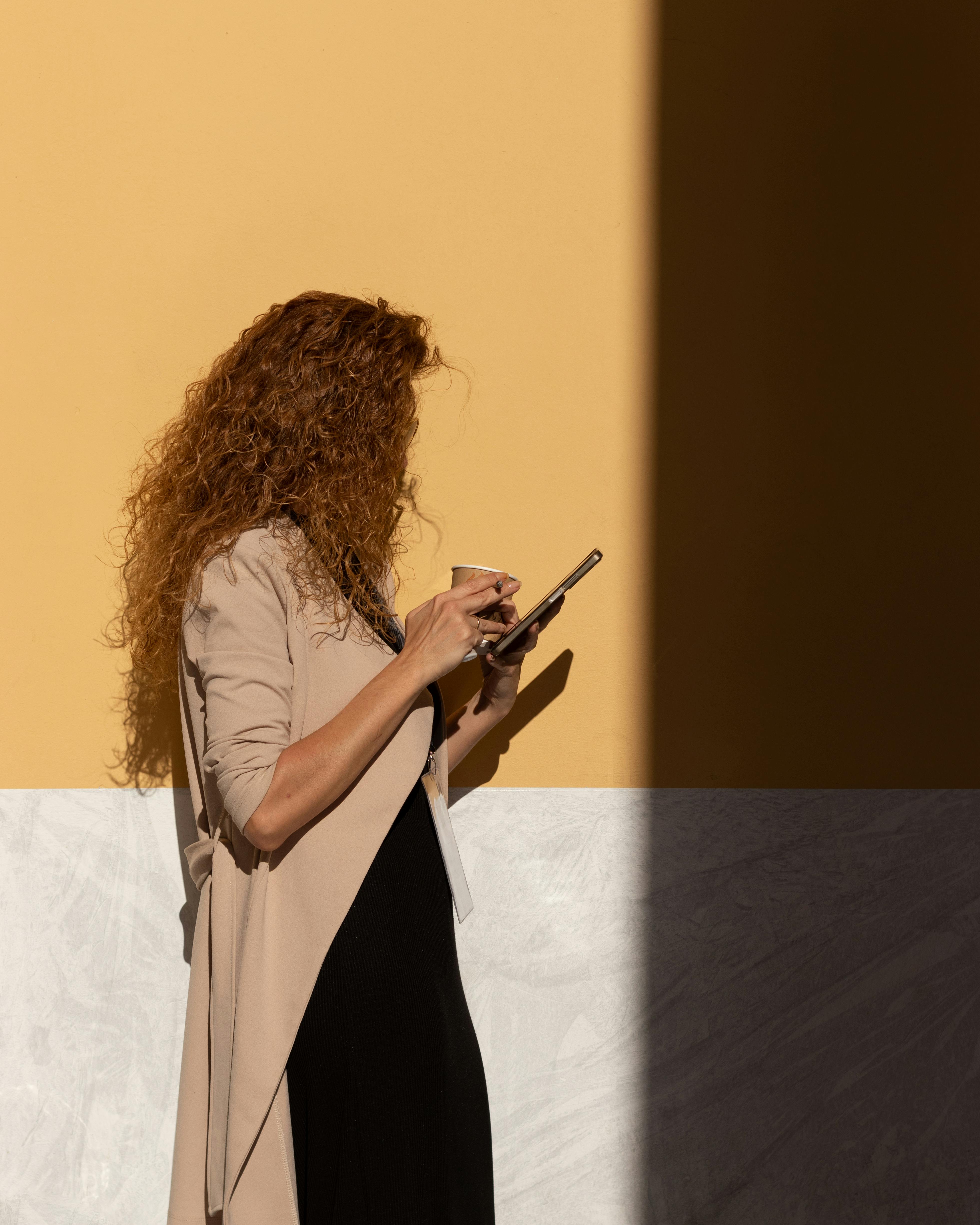 A woman stands by a sunny yellow wall in Athens, Greece, using her smartphone.