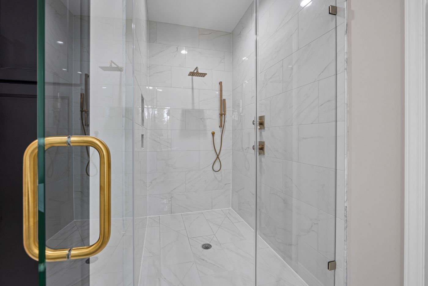 Glass-enclosed walk-in shower with white marble-look wall and floor tiles, gold fixtures, multiple shower heads and a handheld shower.