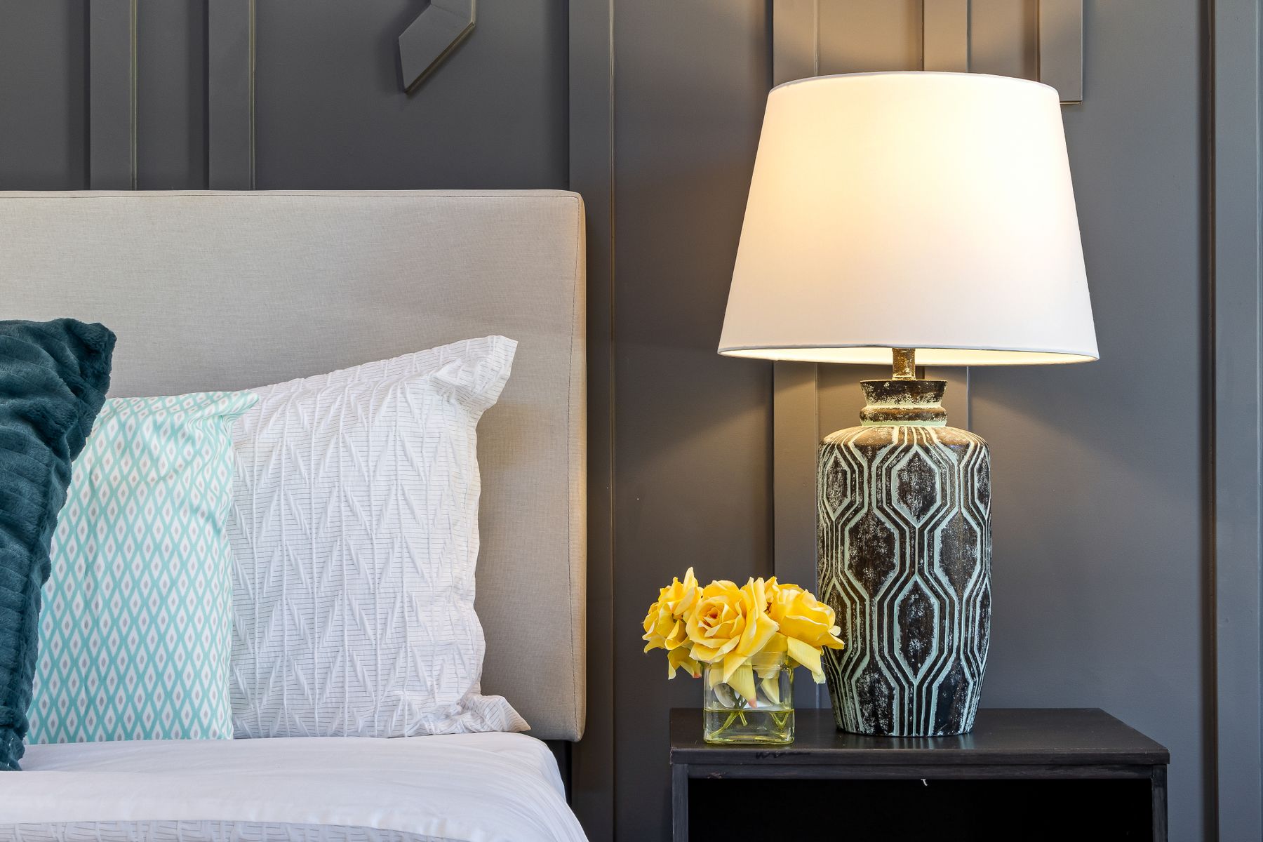 Bedroom detail with warm shaded lamp, brass-tone fixture, pale headboard, white bedding and ochre cushion