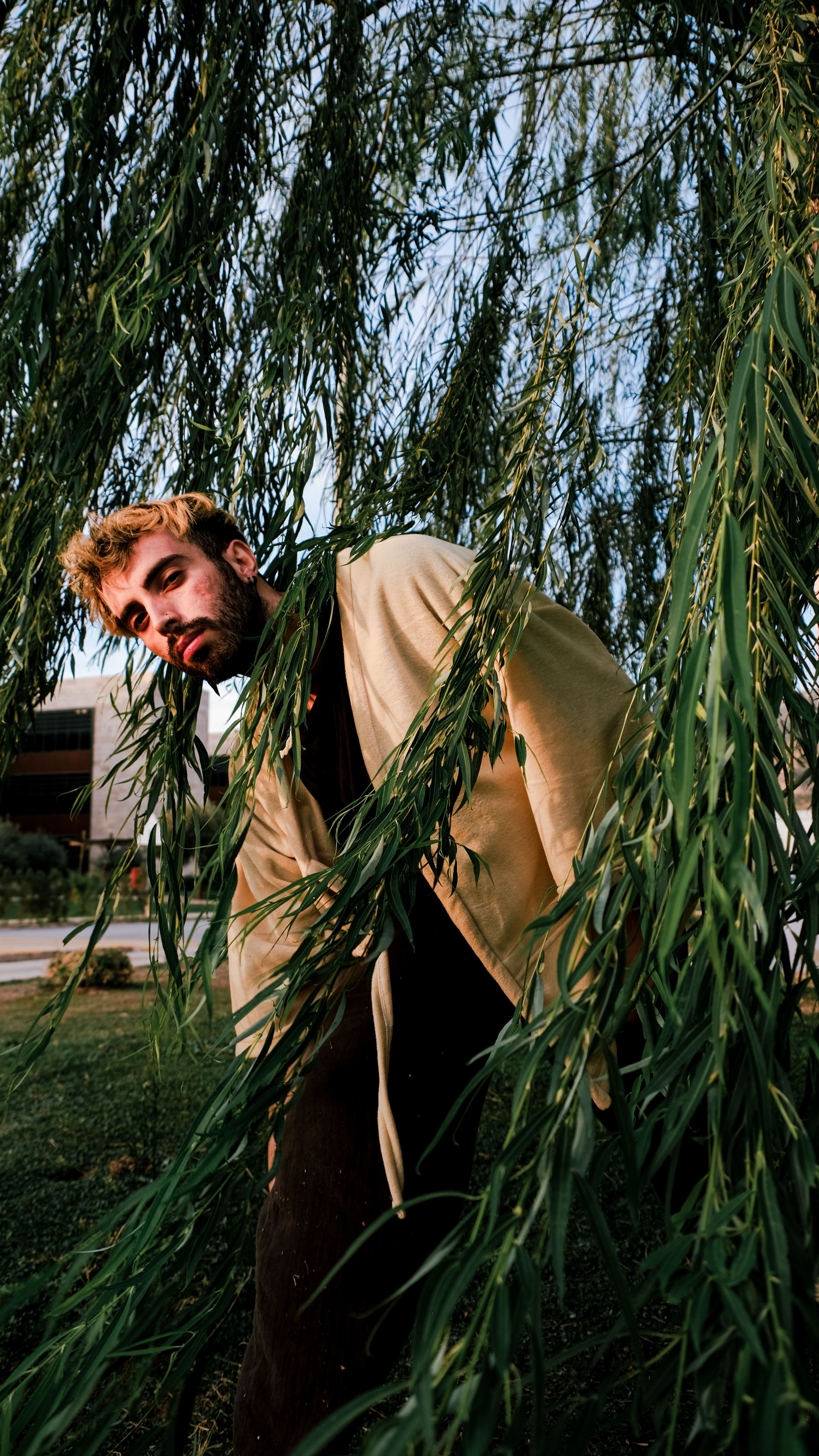 A man poses stylishly among weeping willow branches showing creative fashion.