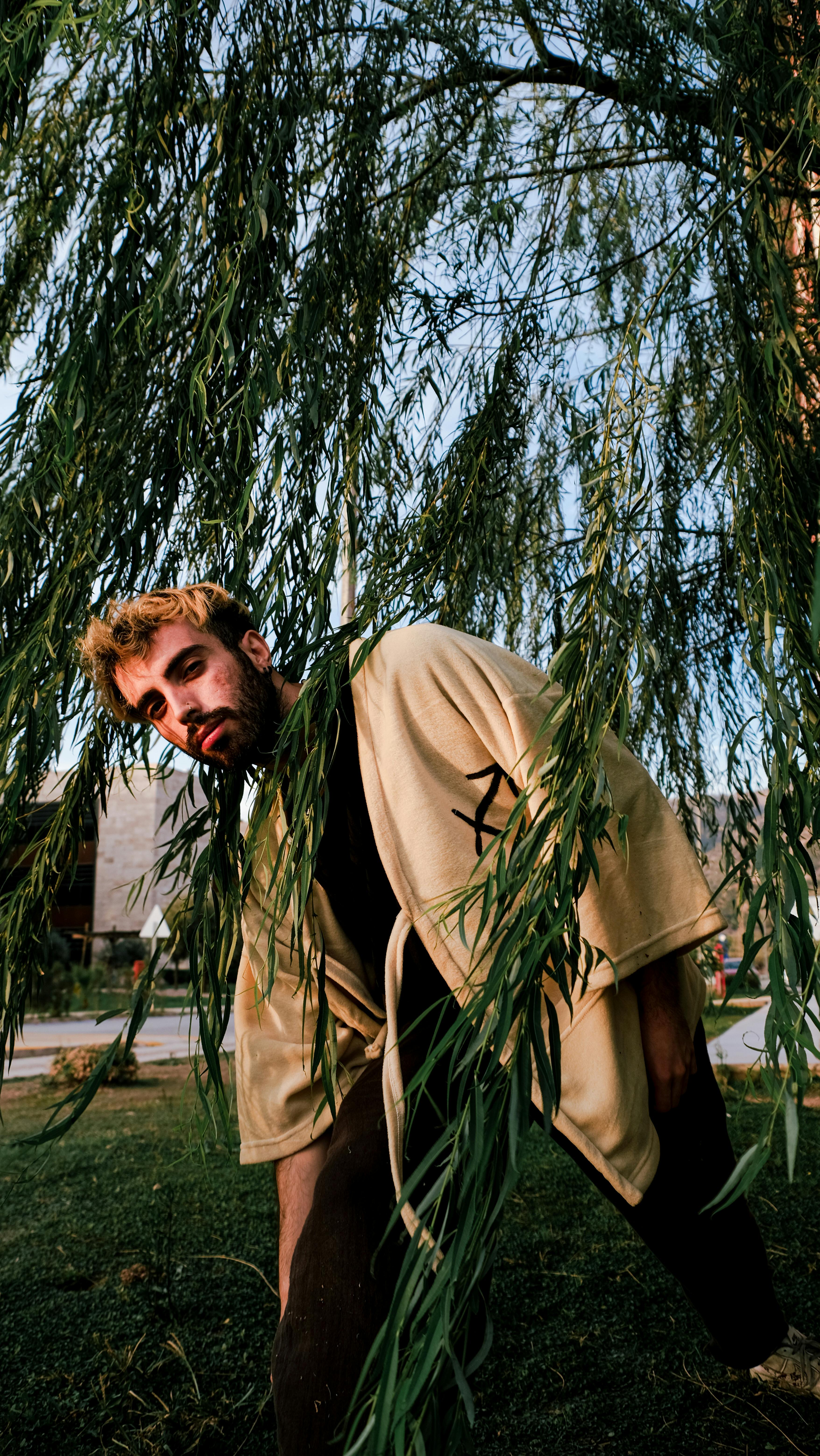 A young man with dyed hair stands amidst weeping willows, creating a natural portrait with a moody vibe.