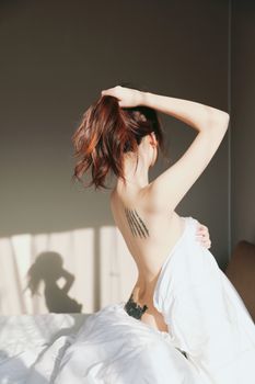 A woman with tattoos sits on a bed, styling her hair in natural light.