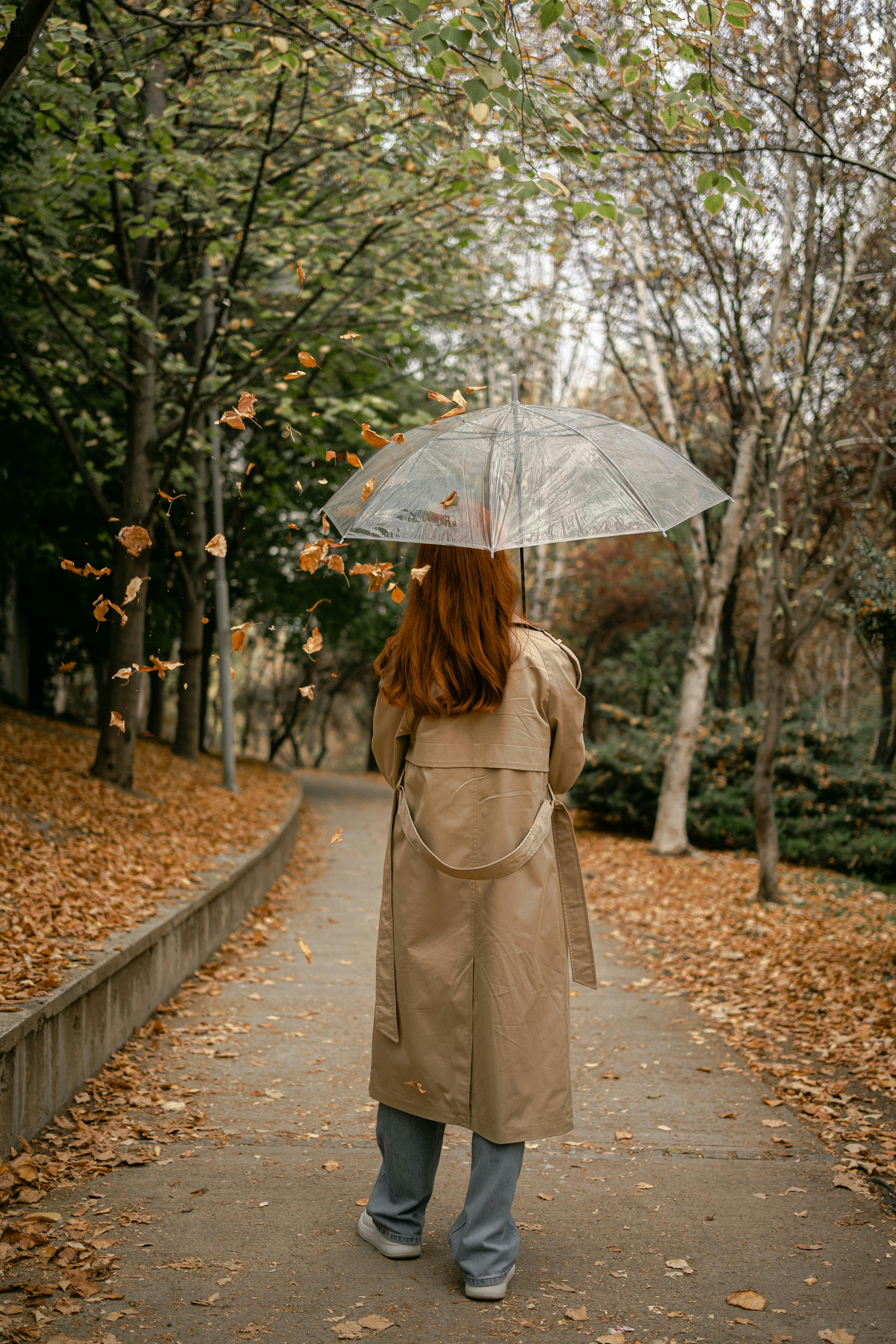 Redheaded woman in trench coat walking under umbrella through autumn park with falling leaves.