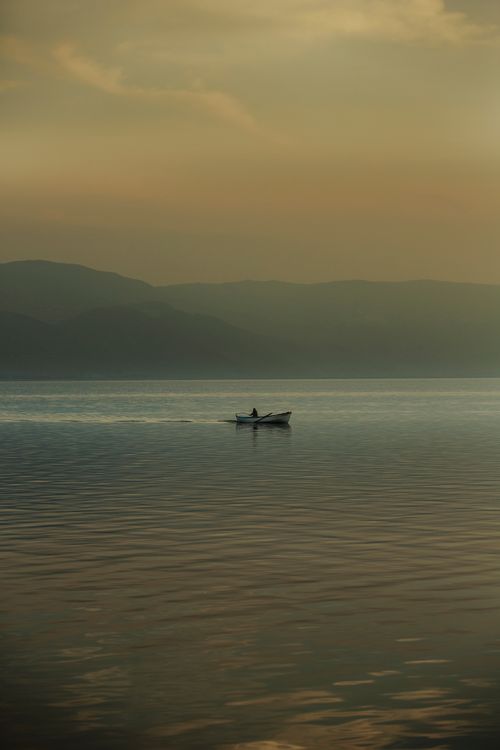 Free A tranquil scene of a lone boat on a misty lake surrounded by hills at sunset, evoking calmness. Stock Photo