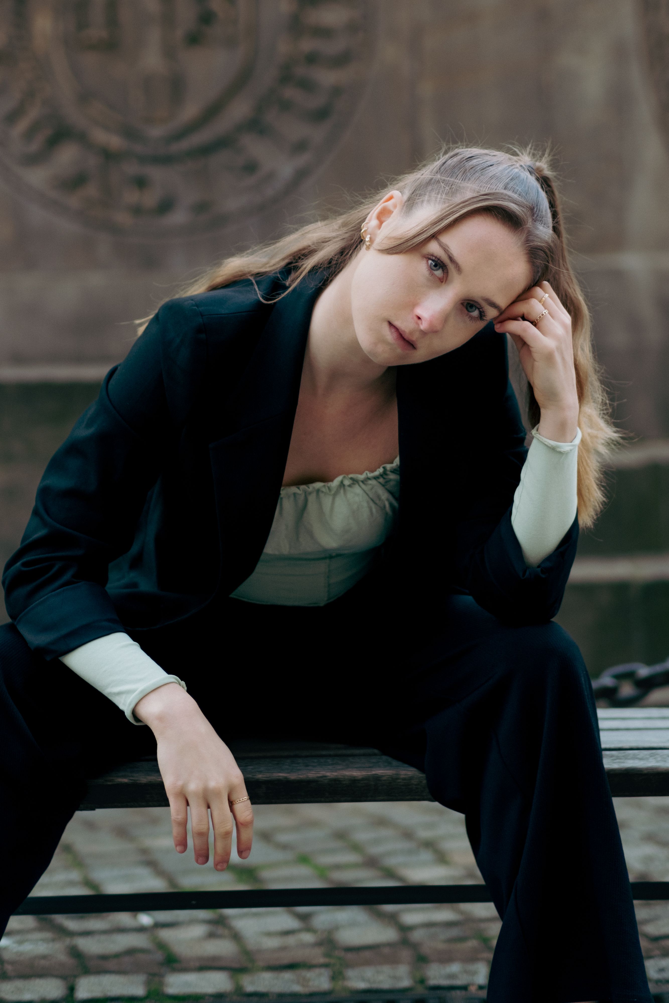 A stylish woman in a black blazer sits thoughtfully outdoors in Berlin, Germany.