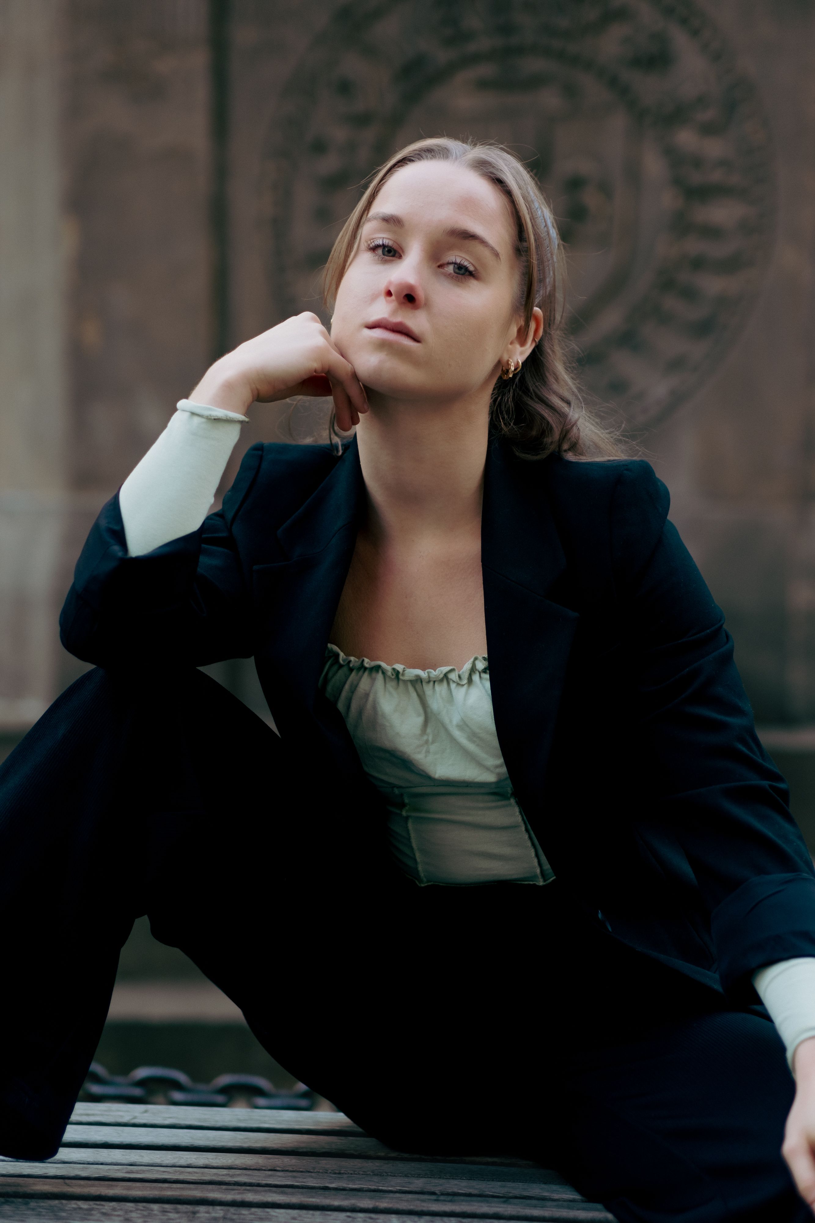 A fashionable woman sitting in a black suit with thoughtful expression in an outdoor setting.