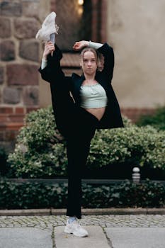 A young woman in sportswear performs a high kick stretch on an outdoor sidewalk.