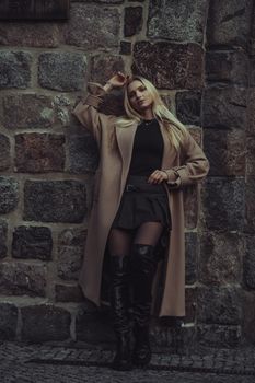 A stylish young woman in Berlin leans against a stone wall, showcasing autumn fashion.