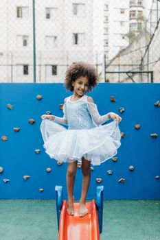 Cheerful girl in a blue dress enjoying playtime on a colorful playground slide.