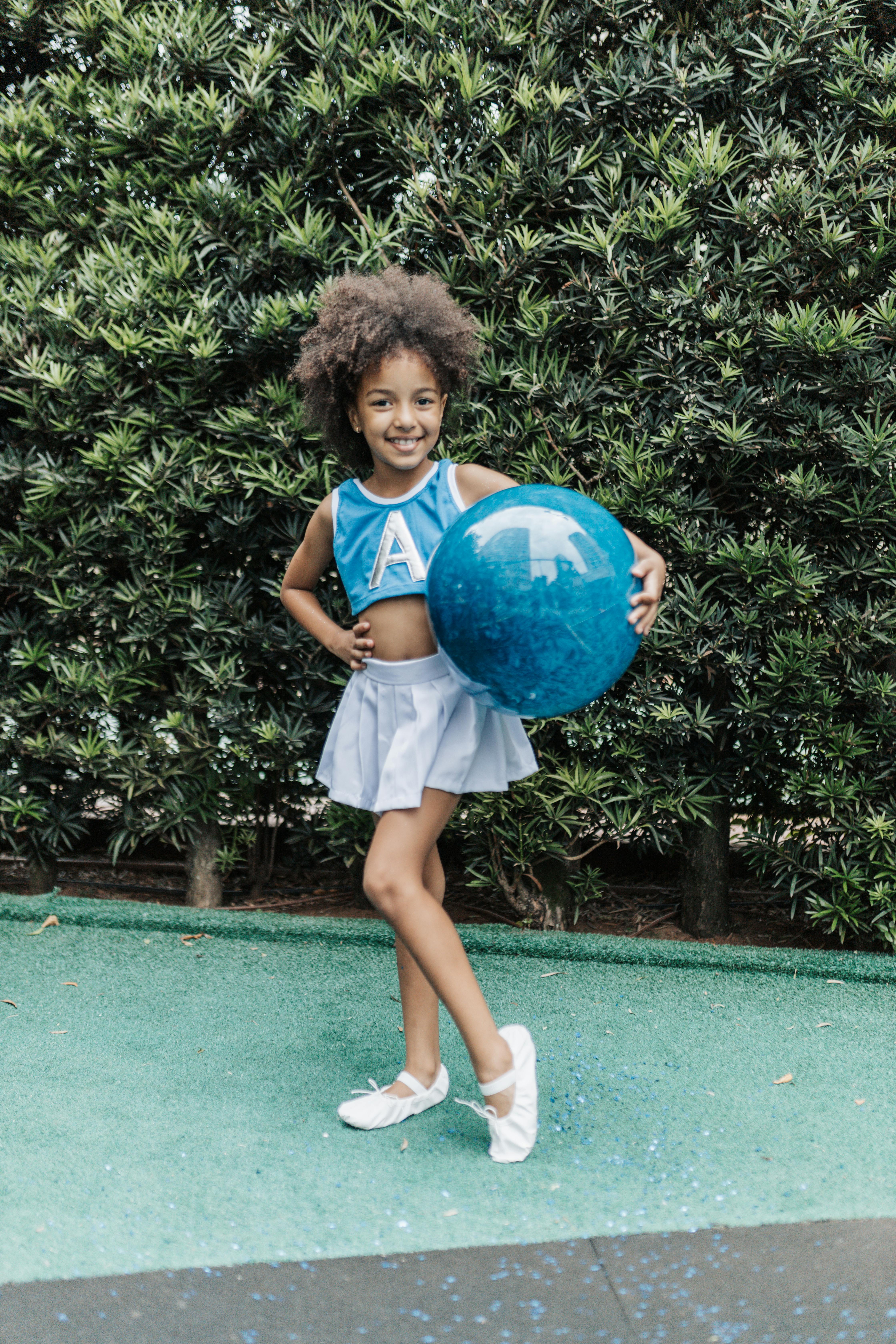 a little girl in a cheerleader outfit and holding a ball standing outside