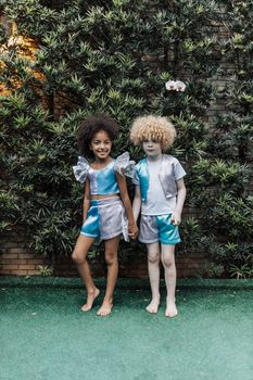 Two children pose outdoors in matching silver and blue outfits, joyful expressions amid greenery.