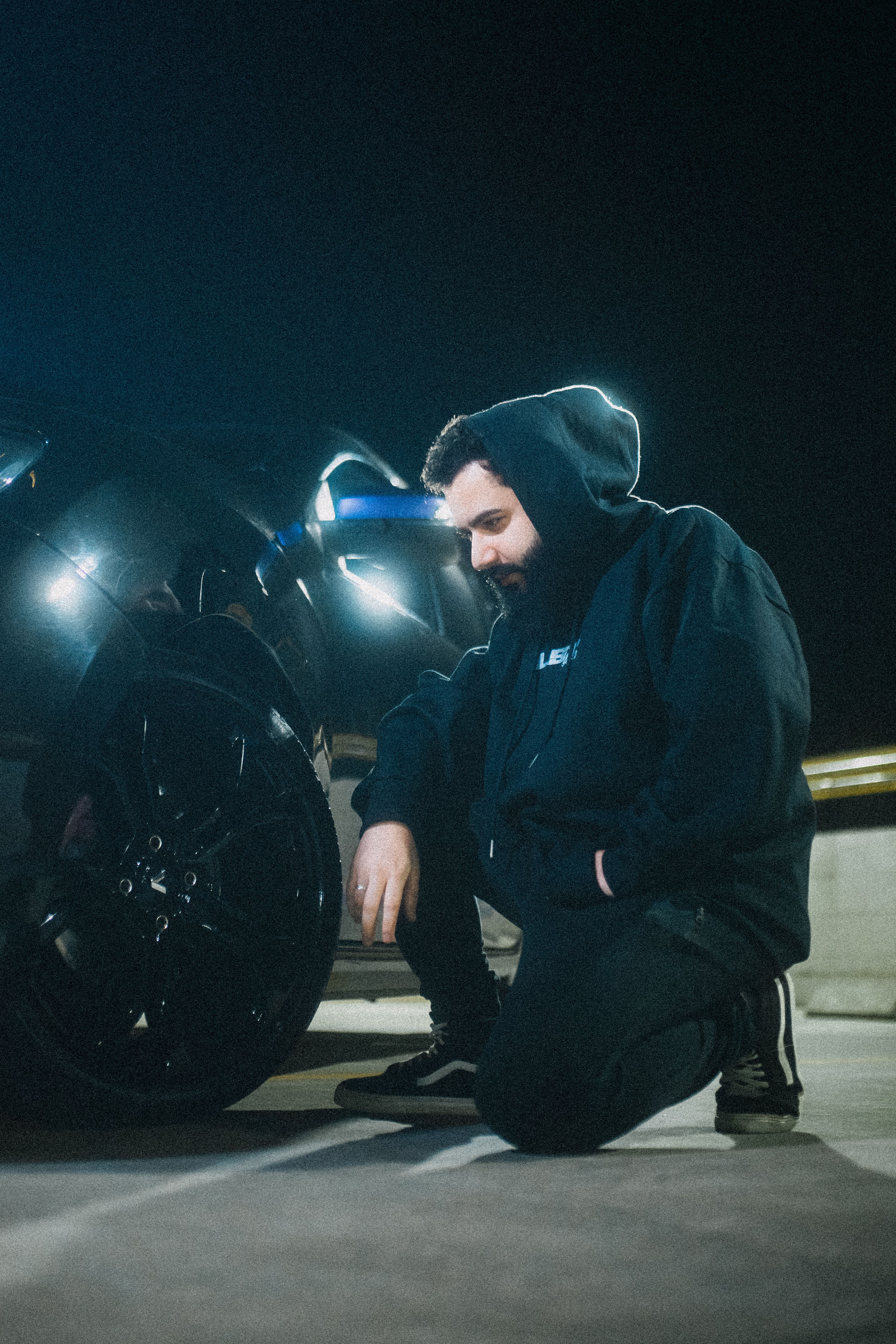A man in a hoodie kneels beside a sleek black car in a dimly lit parking lot at night.