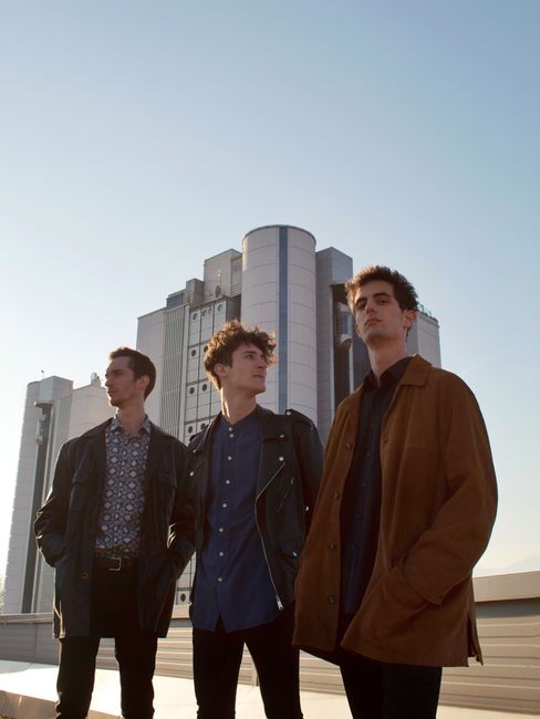 Three fashionable men posing in front of industrial building in Schio, Italy during daylight.