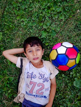 Child lying on green grass beside a vibrant soccer ball, enjoying outdoor leisure.