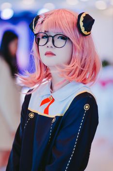 A young girl in anime cosplay outfit with pink hair and glasses, indoors.
