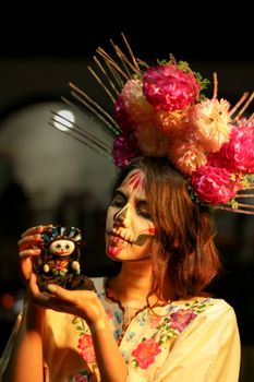 Colorful portrait of a woman in Day of the Dead makeup holding a figurine with traditional floral headdress.