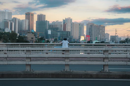 A person stands alone on a bridge overlooking a modern city skyline at sunset.