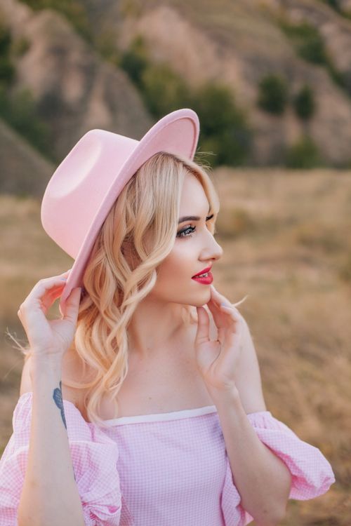 Free Stylish blonde woman in pink outfit and hat posing outdoors with a scenic background. Stock Photo
