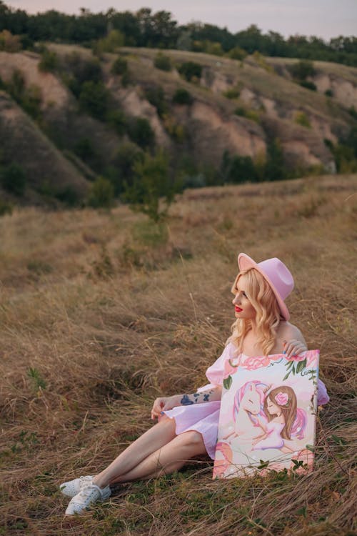 Woman in Pink Dress and Hat Sitting with Painting on Grassland