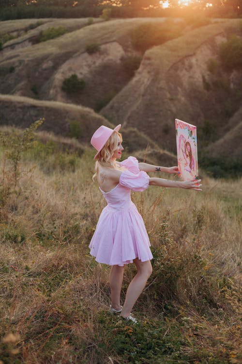 Woman in Pink Dress Standing with Painting on Grassland at Sunset