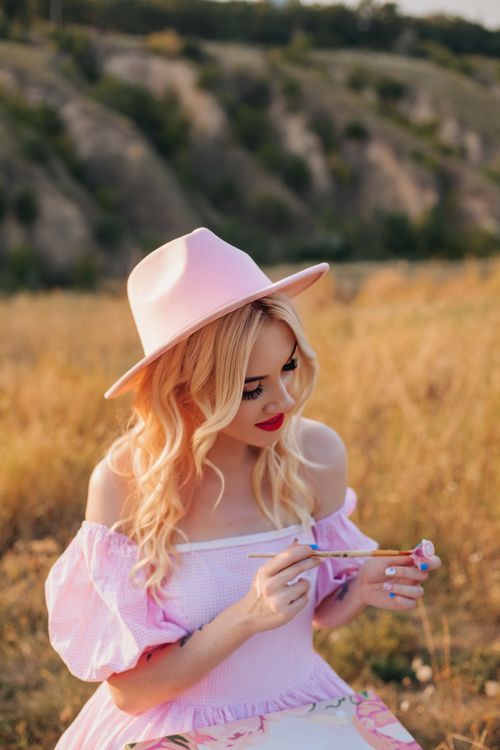 Free Blonde woman in pink dress and hat painting in a scenic landscape. Stock Photo